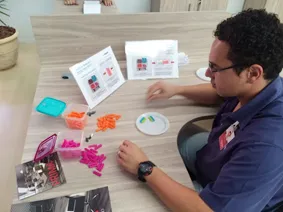 Profissional monta peças plásticas laranja e rosa sobre a mesa, ao lado de fichas ilustradas de instrução
