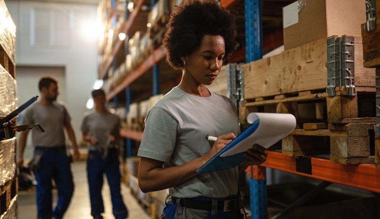 Black female warehouse worker writing delivery schedule list.