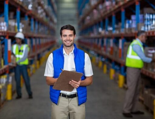 Supervisor de logística em armazem de estocagem conferindo os produtos. O homem está de blusa branca, calça clara e colete azul sorrindo.