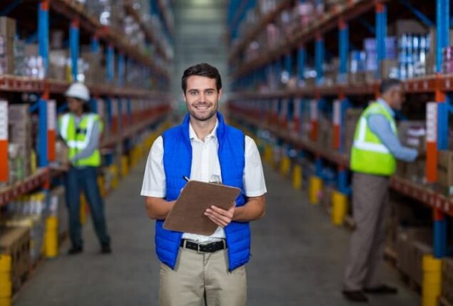 Supervisor de logística em armazem de estocagem conferindo os produtos. O homem está de blusa branca, calça clara e colete azul sorrindo.